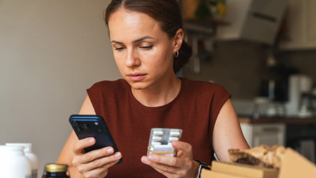 elinde ilaçlar bulunan ve diğer elindeki telefon ile bu ilaçlar hakkında internetten bilgi sahibi olmaya çalışan kadın görseli